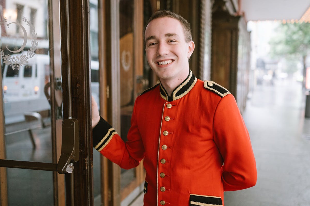 An usher in a red coat holds open the foyer doors for a patron