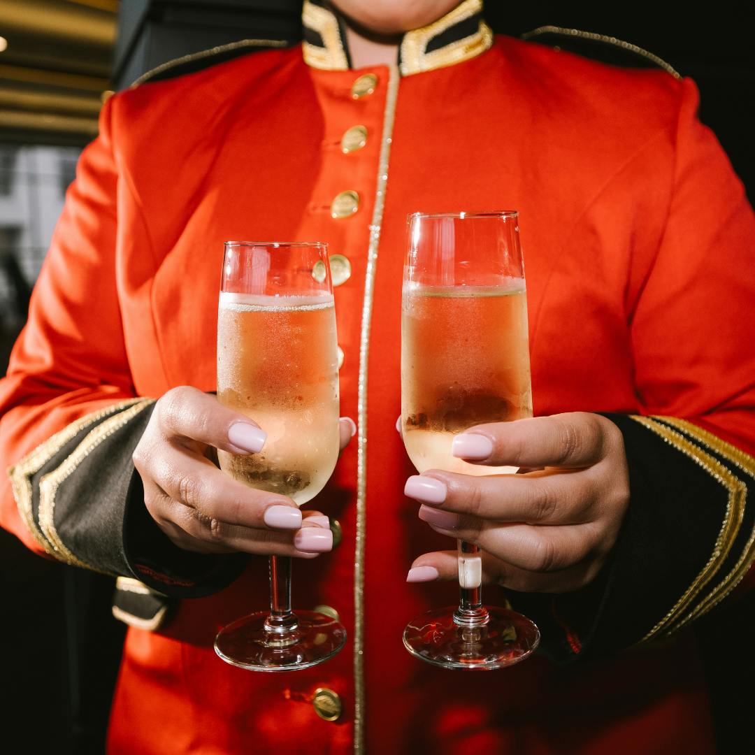 An usher wearing a red coat holds two glasses of champagne. Their face is not visible.